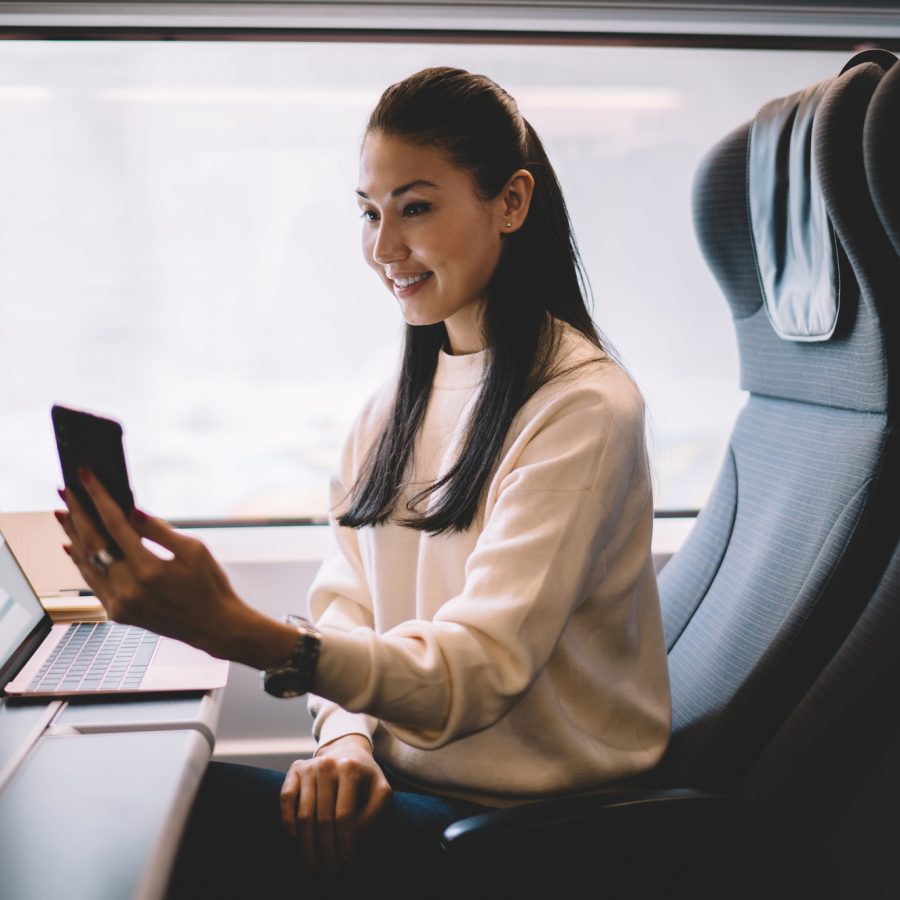Cheerful young Asian woman in casual clothes sitting at table with laptop and coffee and smiling while taking selfie on cellphone in contemporary train